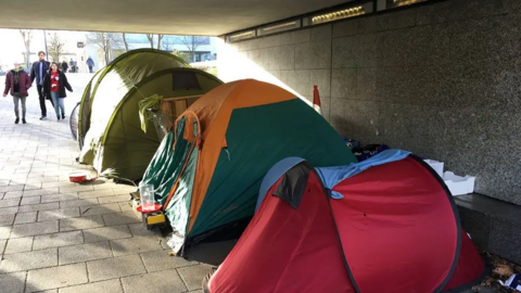 Several camping tents are pitched under a pedestrian underpass beside a dark stone wall. The tents, in red, green and orange, line the sheltered walkway, with bedding and personal belongings visible nearby. A few pedestrians walk past in the background where sunlight enters the underpass.