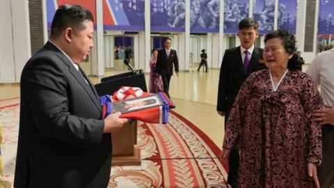 Kim Jong Un, wearing a dark suit, presents a crying woman with a photo draped in a blue and red flag. The woman is wearing a dark red dress and they are in a grand hall. 