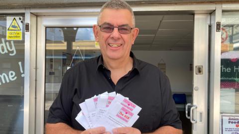 A man wearing a black shirt is standing in front of an office building, holding up several sheets of paper which have ideas written on them.