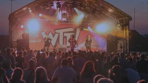 The festival in Melksham in 2025. A three-piece musical group, two in sparkly black clothes and one in a sparkly pink outfit, performing on a stage at night time. Behind them is a large sign reading "WTF" – referring to Wiltshire Throwback Festival. Outlines of audience members are in the foreground.