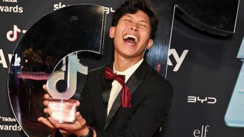 Gab Astorga, who has black wavy hair, closes his eyes, throws his head back and laughs , while holding a glass and mirror TikTok award. He is dressed in a black dinner suit with red bow tie and is standing in front of a black branded screen.