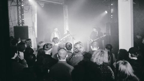 A live show inside The White Hotel venue. Two men are playing guitars on stage and a crowd watch.