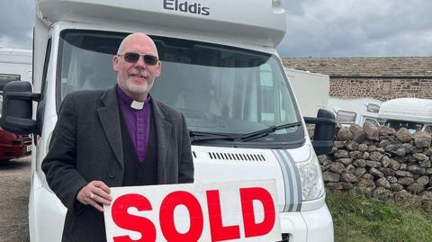 Bishop Mick Fleming wearing a purple shirt with a white clerical collar holds a sign saying 'sold' in red with a motorhome behind him. He is smiling. There are a number of other motorhomes in the background behind a stone wall and a farmhouse. 