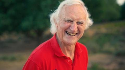 A man with white hair smiles widely at the camera while walking outdoors. He is wearing a vibrant red collared t-shirt.