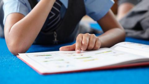 A child with a pale blue shirt, black waistcoat and stripy blue and white tie has a finger on an opened page of a book with their other arm's elbow resting on a blue surface. You cannot see above their shoulders.