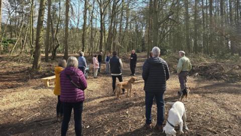 A group of dog walkers wearing coats and with dogs on leads stand on a path in a wooded area.