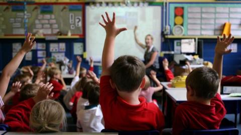 Stock image of a class full of primary pupils raising their hands, with a teacher pointing to a whiteboard.