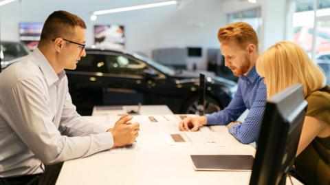 Salesperson wearing a white shirt advising couple in car dealership as they sit around a table, with a black car in the background