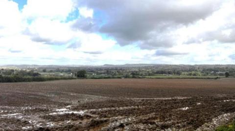 A view over a muddy field with some houses in the distance.