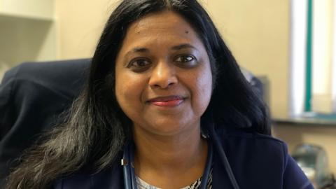 Dr Kuttappan smiles at the camera with a stethoscope around her neck. She has long dark hair and is wearing a blue jacket with a coat hanging on the back of her chair