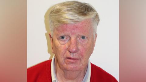 Mugshot of a man with a red jumper and grey hair looking at camera.