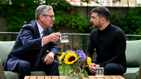 Prime Minister Sir Keir Starmer (left) and Ukrainian President Volodymyr Zelensky in the garden of number 10 Downing Street. They sit on a green sofa in front of a wooden table with a glass vase filled with yellow sunflowers and blue flowers, representing the colours of the Ukrainian flag. They have matching Downing Street mugs and Sir Keir is raising his as he talks to Zelensky with serious expressions on their faces.