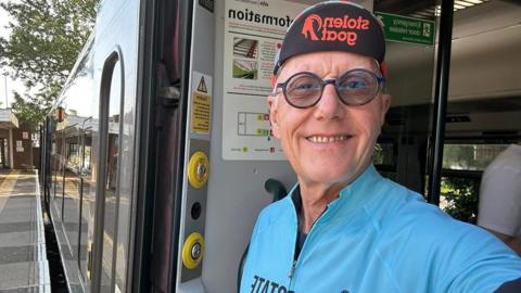 A man wearing a cycling jersey stands in the open doorway of a train at a station platform, with the train visible beside them.
