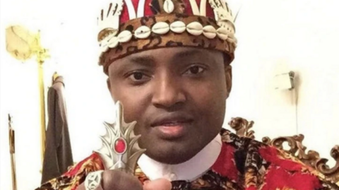 Simon Ekpa wearing a crown decorated with shells and holding a staff