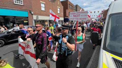 A demonstration waving England flags walking down a high street lined by brick buildings. One placard is visible, which reads "Illegal immigrants are not welcome. Stop the boats."