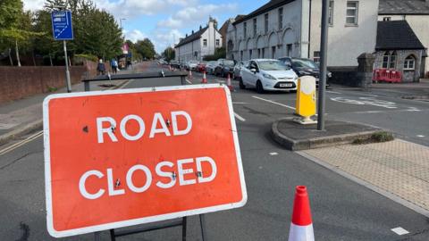 A large red and white road closed sign in block capitals at the junction of Gladstone Road and Heavitree Road in Exeter with cars queuing in the opposite direction.