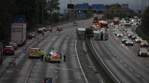 A motorway with a white lorry on its side straddling the central reservation. Police cars in the foreground