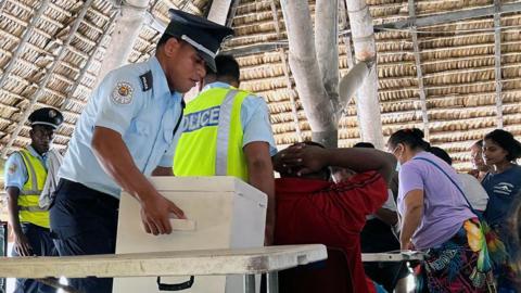 A police officer handles a ballot box as people sit at tables under a thatchwork ceiling.