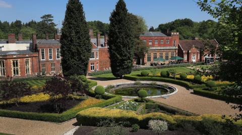 A gravel path leads to and around a water feature in a manicured garden, with carefully pruned hedges and trees. The path leads to a large brick manor house.