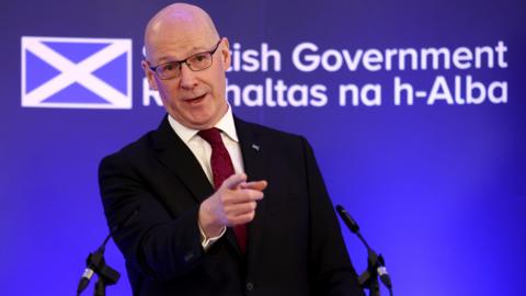 First Minister of Scotland John Swinney points at the audience while making his speech at the Royal Society of Edinburgh. He is standing at a lectern against a bright blue backdrop emblazoned with a Scottish government logo and a saltire. Swinney is wearing a black suit, a white shirt and a red tie.