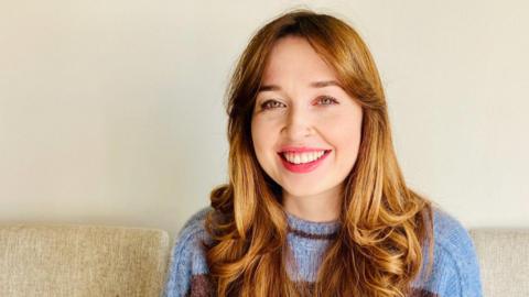 Kate Driver is smiling at the camera. She has slightly curled hair and is wearing a light blue and purple jumper.