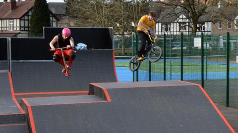 A man with a pink helmet on a scooter and a man on a BMX get some big air as they jump a ramp on the new skate park. 