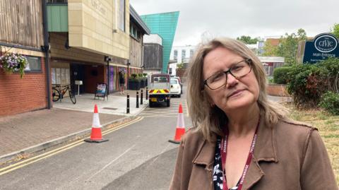 A middle-aged women holds her head at an angle while looking sternly at the camera. She is wearing a glasses, brown jacket, a maroon lanyard and there are orange cones, buildings, vegetation and a sign with the word "Civic" written on it behind her.