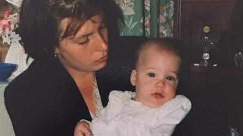 A woman with short dark hair holds a baby in a white dress.