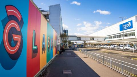 A colourful sign at Glasgow Airport. The airport on the right is a long, white building with white taxis parked outside it.
