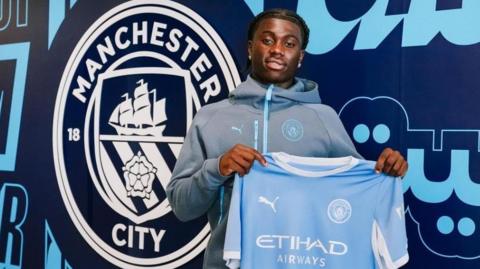 Jeremy Monga holding up a Man City shirt.