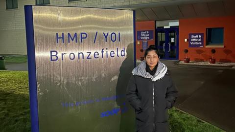 A woman in a long coat and black and white check scarf, standing outside a prison with her hands in her pockets