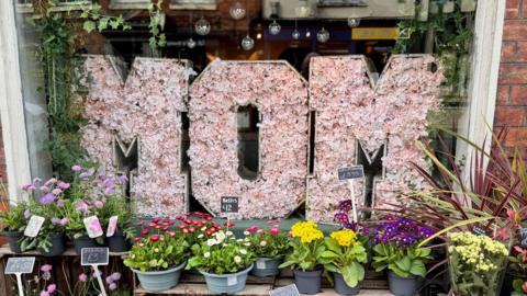 A floral display in the window of a flower shop spells out the word MOM in pink flowers. In front of it are pots of different coloured flowers and grasses, each individually priced.