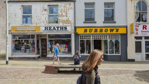Shops on a street in Barrow town centre. One has a Toymaster sign above the window, while to its right is Heaths Stationers & Booksellers. Much of the paintwork on the building is peeling. Two people are walking along while a third is seated on a wooden bench.
