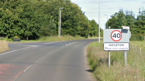 A country road with grassy fields on either side with trees in the distance. A sign says the speed limit is changing to 40mph and it also reads "Houston, please drive safely"