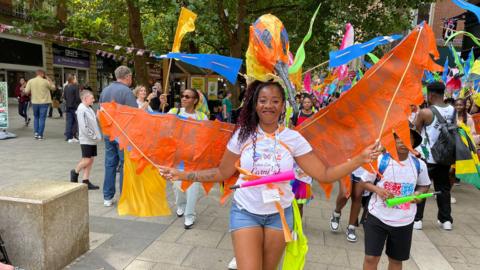 A woman holding out a carnival costume, made of orange wings, wearing a white T-shirt and blue shorts. Lots of people are behind her in the middle of a busy city centre. 