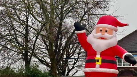 A giant inflatable Father Christmas on the right with trees to the left and grey sky behind