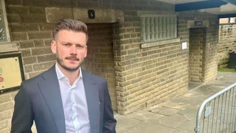 A man with short dark hair wearing a white shirt and grey suit jacket stood outside a public toilet