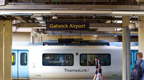 Sign at Gatwick Airport railway station with platform and a white Thameslink train departing the station. People are stood on the platform.
