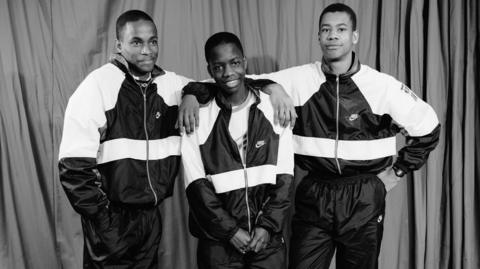 Boys from Tulse Hill School, 1990. From left: Mark De-Lisser, Edmund Rudder and Marcellous Copeland. The trio stand in front of a curtain. They wear matching Nike tracksuits and trainers. Black and white photo.