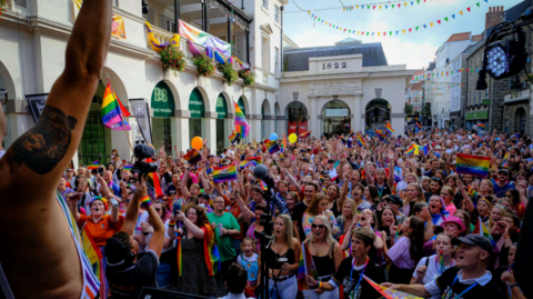 A large crowd of people holding up various flags. There are buildings to the left and right of the crowds. There is rainbow bunting. The sky is blue with white clouds.