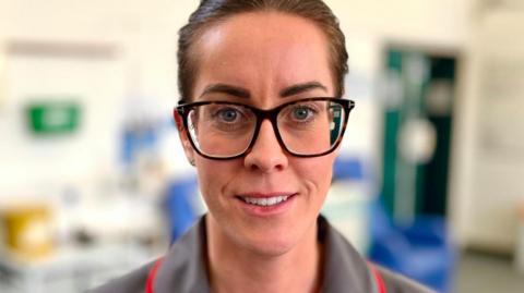 Matron Lana Goodwin has brown glasses on, has her hair tied back and has a grey uniform with red seams. She is looking directly at the camera, she is standing in the Billericay Covid clinic. 