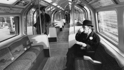 A black and white image of a London Underground official drinking a cup of coffee during trials of new automatic trains on a section of the Central Line.