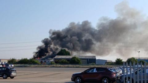 A large plume of black smoke coming from an industrial building in the distance. The photo is taken from a car park where few cars are parked. The sky is blue.
