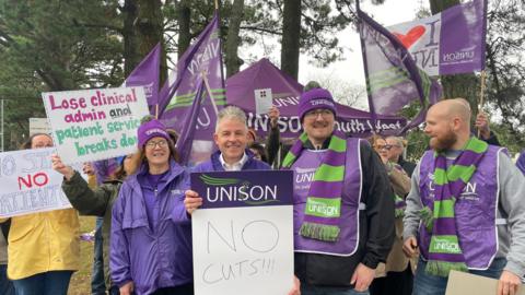 About nine people, in purple and green clothing, holding placards. Some people in the back are holding UNISON and "I LOVE NHS" flags. There are trees in the background. The sky is grey.