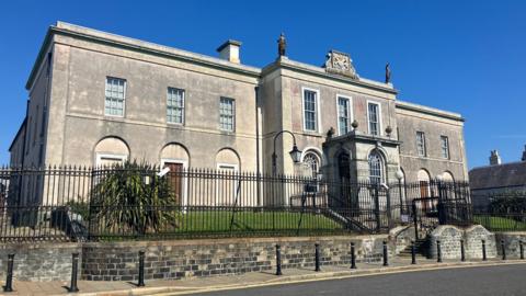 A large grey building stands on top of a green bank, with steps leading up to its main entrance. The building is surrounded by a tall black-metal fence. Ther sky above is clear and blue. The building sits beside a black tarmac road. A grey footpath seperates the road from the building.