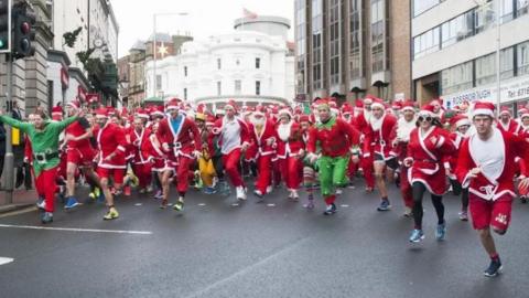 Runners dressed in Santa outfits and elf costumes set off on  Prospect Hill. A man on the left wearing a green elf top and red trousers has his arms stretched out to the sides as he sets off. The round white Tynwald Wedding Cake building can be seen behind them.