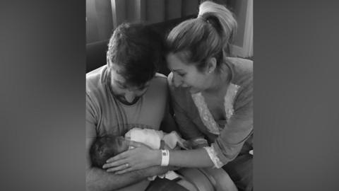 Black and white photo of Dan and Fiona cradling their newborn baby. Dan holds her in his arms and is looking down at her, while Fiona strokes her face. A hospital band is visible on her wrist.