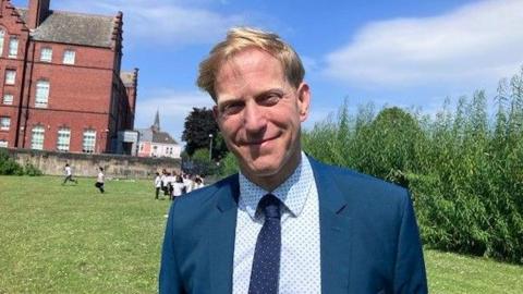 Karl Ellerbrook smiling into the camera. He has short blonde hair and is wearing a blue suit with a white patterned shirt and blue patterned tie. He is standing on a grassy field with children behind him, a couple of them are running to the left. Behind them is a large, red brick Victorian school building. The sky is blue with a few clouds.