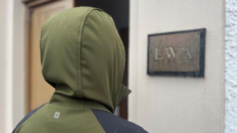 Unidentifiable person with their back to the camera wearing a khaki waterproof jacket with the hood up. They are standing outside a building with a sign that says "LWA".