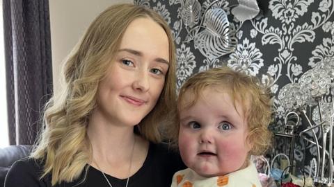 A young woman in her early 20s with wavy, long blonde hair and wearing a black top and silver heart pendant necklace is holding a baby of 13 months. He is wearing a baby grow with a pumpkin pattern and has reddish blonde curly hair and rosy cheeks. The woman is smiling at the camera and the baby is smiling looking just off to the side. There is black and silver wall paper behind them and a grey curtain hanging at the window.